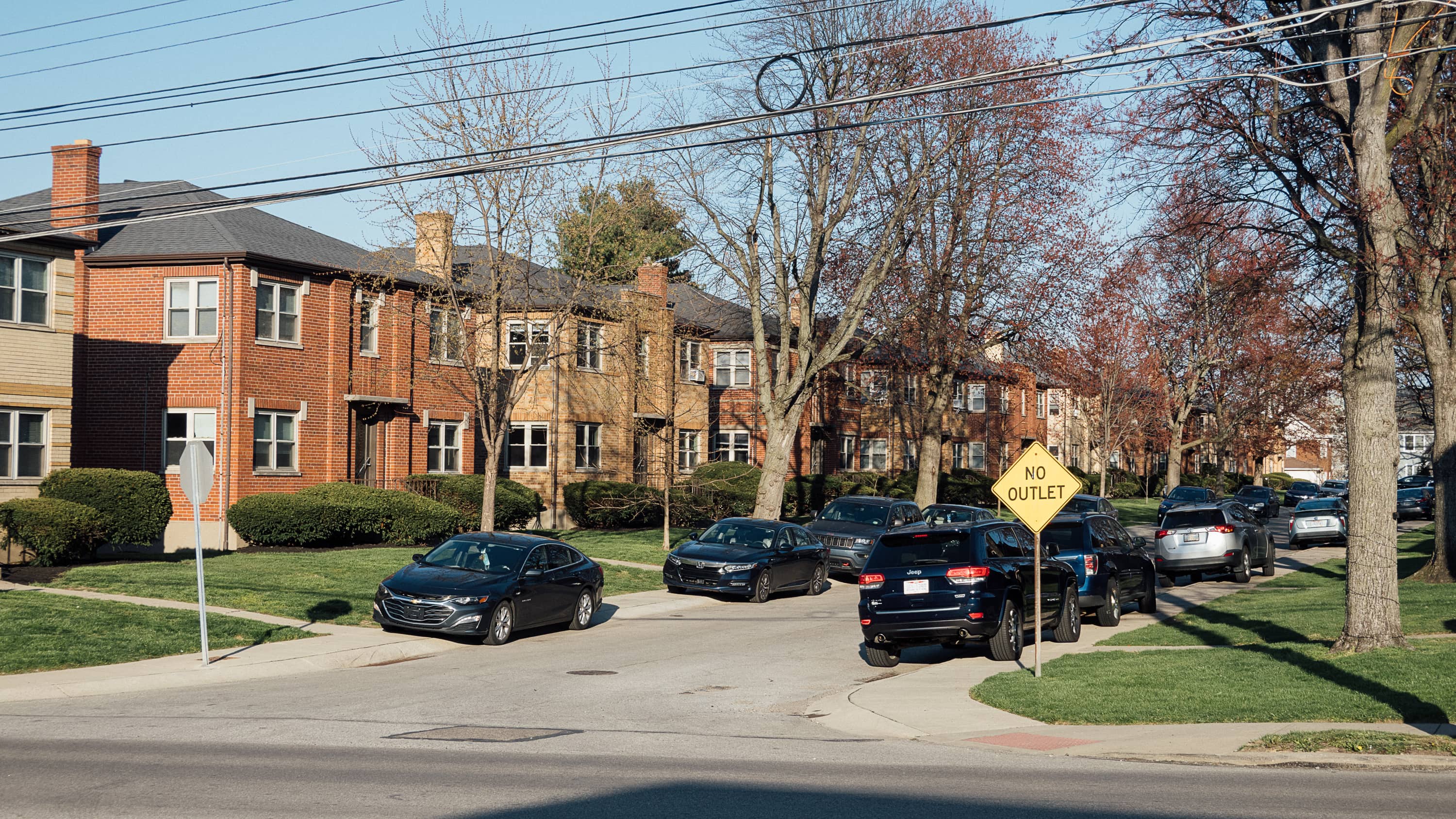 A dead end street filled with quad apartments and all the cars parked on the street are halfway on the sidewalks on both sides